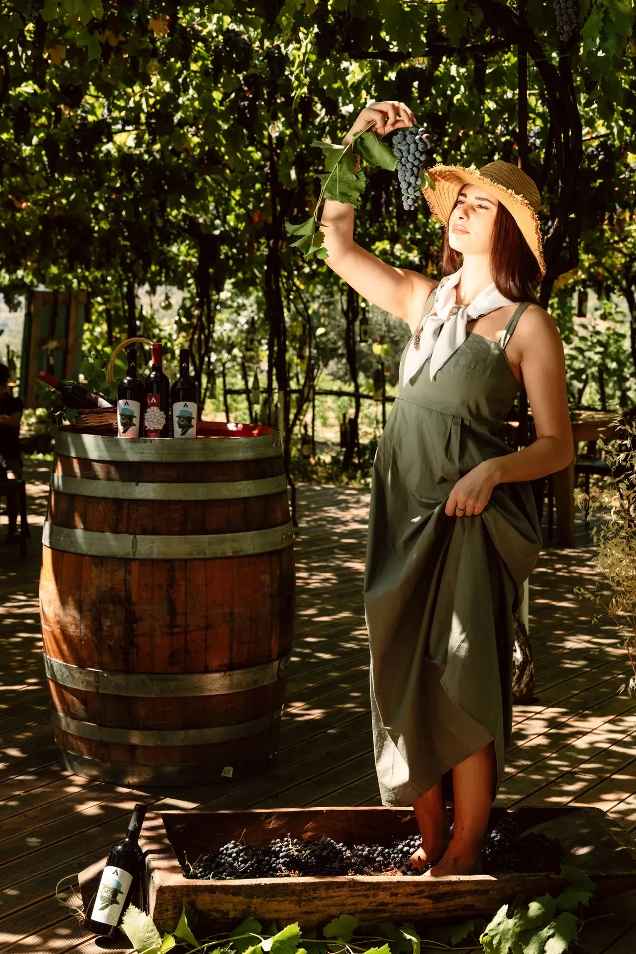 A woman in a straw hat treading grapes in a wooden trough under the pergola, bottles of Xha Beqo on a barrel beside her