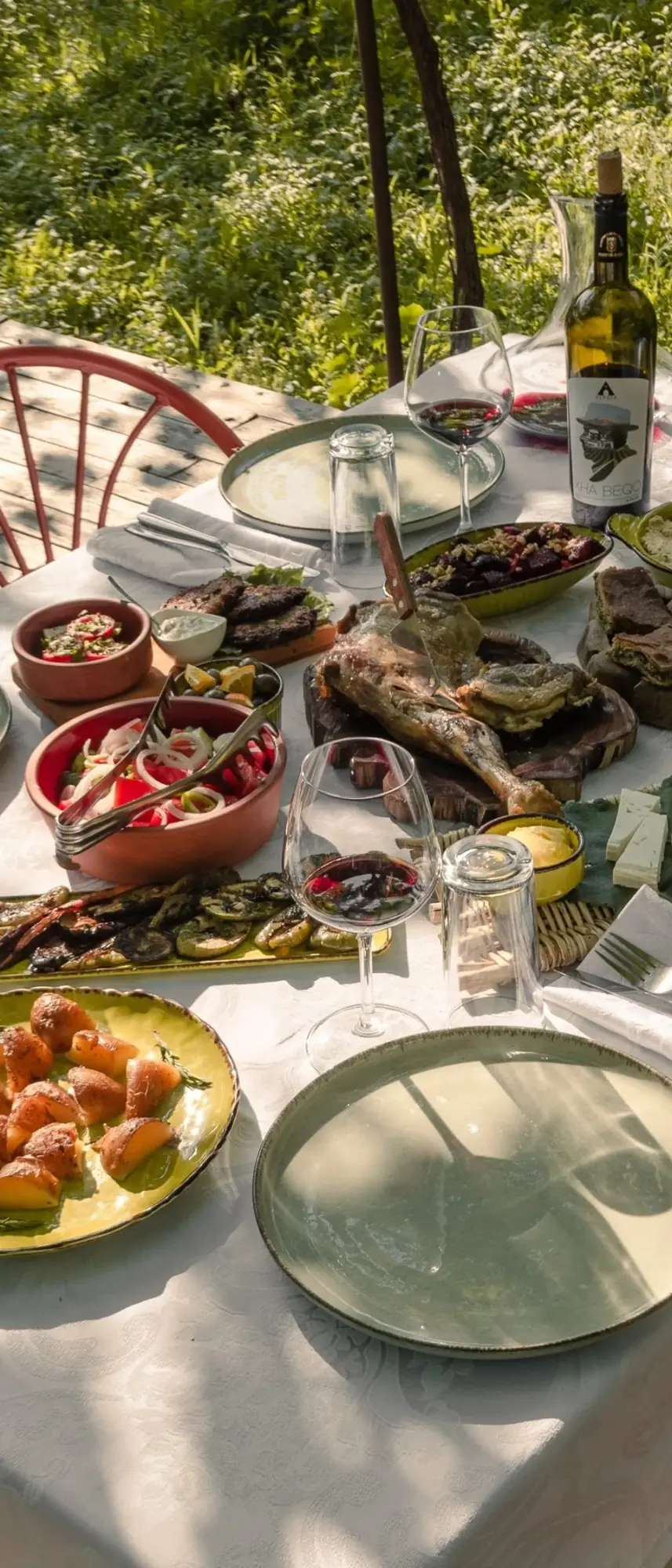 A long garden table in the sun laid with roast lamb, salads, byrek, cheese and glasses of Xha Beqo red wine