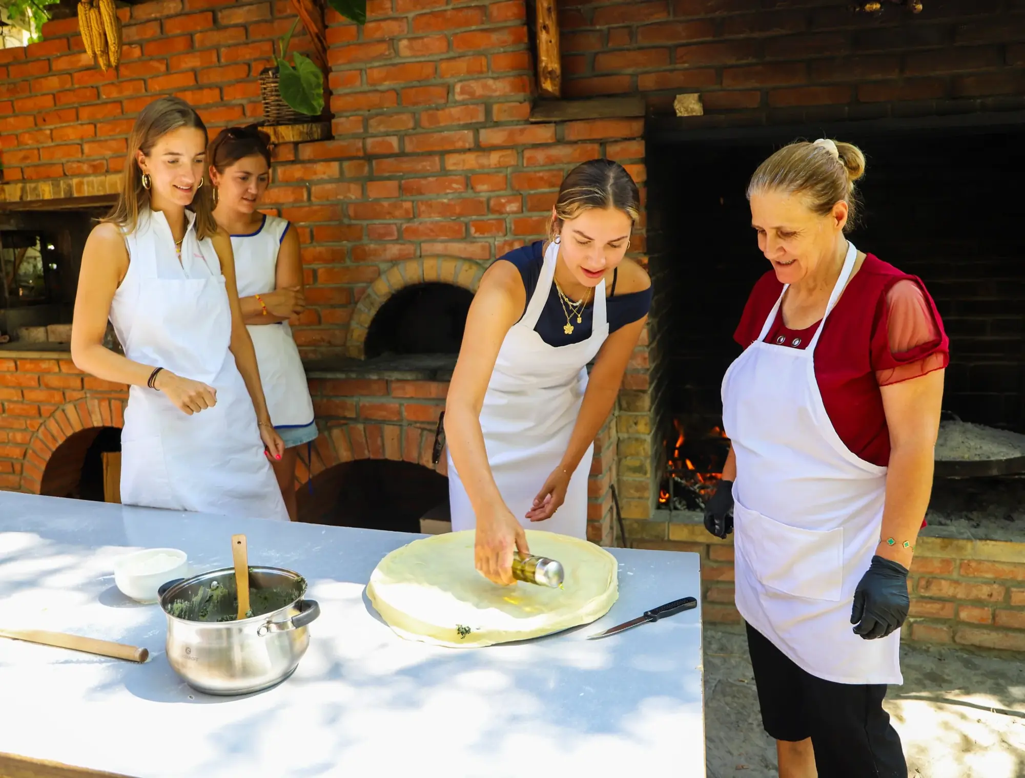 Guests rolling out pastry and oiling it with a local cook beside the brick outdoor oven and open fire