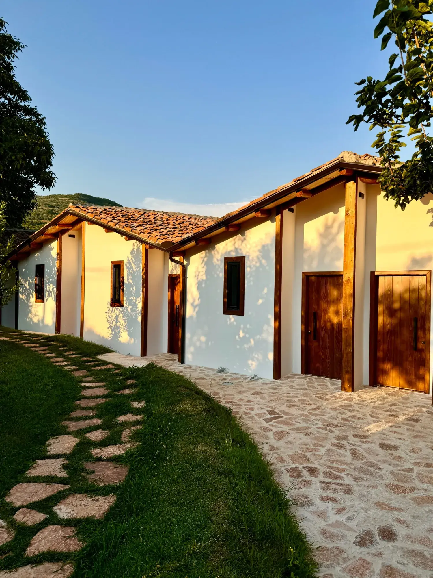 The guesthouse in evening sun: white walls, oak beams, wooden doors and a stone path through the grass, hills behind