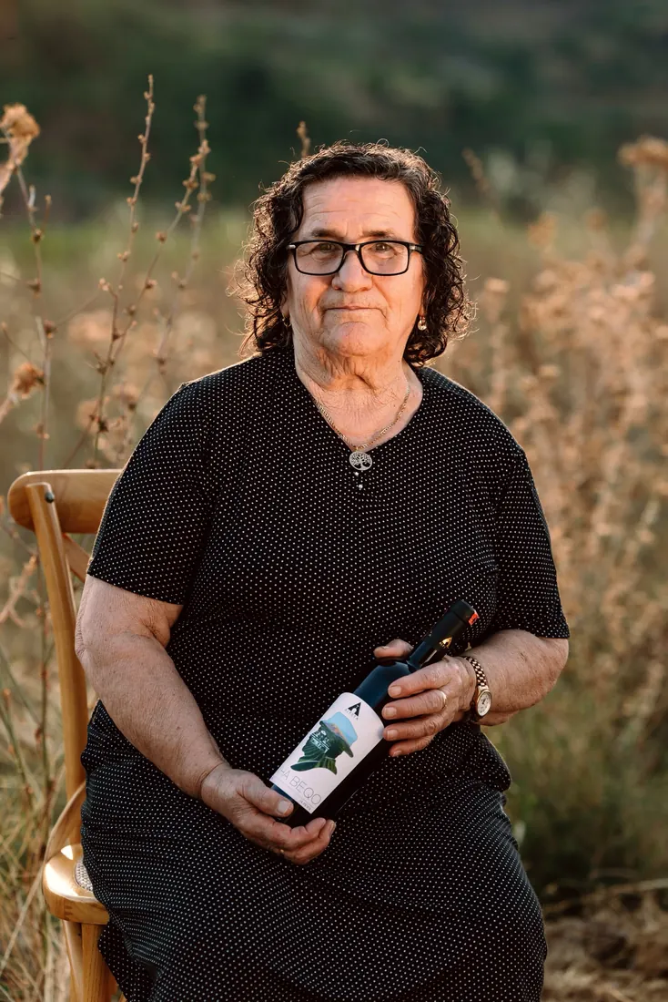 An older woman sitting in the dry grass, holding a bottle of Xha Beqo